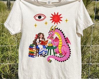 May include: An ivory short-sleeve t-shirt featuring a graphic print of a person with long brown hair reading a book next to a stack of colorful books and a pink dragon. The design includes a yellow teapot, a small green table, and celestial symbols.
