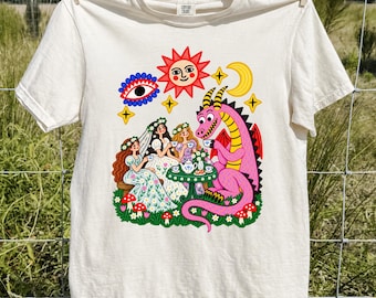May include: An ivory t-shirt featuring a colorful graphic print of two people in floral dresses and a pink dragon having a tea party. The design includes a red sun, a yellow crescent moon, a blue eye, stars, and red mushrooms on green grass.