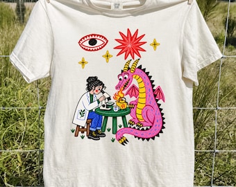 May include: An ivory short-sleeve t-shirt featuring a graphic print of a scientist in a white lab coat using a microscope at a green table, while a pink dragon breathes fire onto a yellow kettle. Above are a red eye, a red star, and yellow stars.