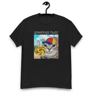 Op de afbeelding: Zwart T-shirt met de tekst "greetings huzz" en een afbeelding van een kat met een kleurrijke hoed en een lolly.