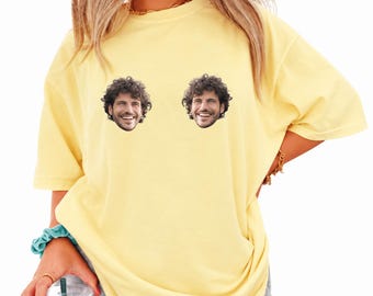 May include: A pale yellow short-sleeved t-shirt featuring two identical photographic prints of a smiling person with curly dark hair positioned on the upper chest area. The garment has a crew neckline and a loose, relaxed fit throughout the body.