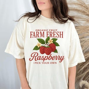 May include: Cream-colored t-shirt with a raspberry graphic. The design includes the text "Organic Fruit Farm Fresh Raspberry Pick Your Own" in a vintage style. The graphic features three red raspberries, green leaves, and small yellow flowers.