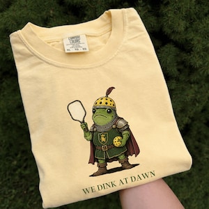 Könnte beinhalten: Ein blassgelbes T-Shirt mit einer Grafik eines grünen Frosches in mittelalterlicher Rüstung, der einen Pickleball-Schläger und einen gelben Pickleball hält. Der Text unter dem Frosch lautet WE DINK AT DAWN. Das T-Shirt hat ein Comfort Colors Etikett.