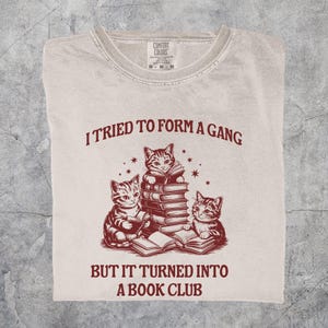 May include: A light beige t-shirt with a crew neck, featuring a maroon graphic of three cats reading books. The text above reads "I TRIED TO FORM A GANG" and below, "BUT IT TURNED INTO A BOOK CLUB."