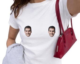 May include: A white short-sleeved t-shirt featuring two identical photographic prints of a smiling man's face positioned on the chest area. The garment is paired with a red shoulder bag and a light gray fabric item held in the lower section.