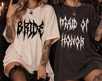 May include: A white t-shirt with the word BRIDE printed in a black gothic font and a charcoal gray t-shirt with the words MAID OF HONOR printed in a white gothic font. Both shirts feature short sleeves and a crew neckline in a relaxed fit.