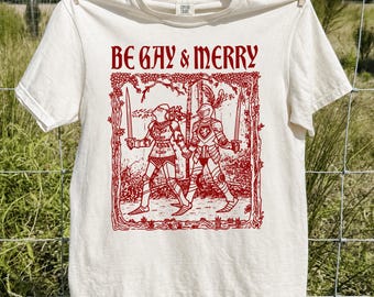 May include: An ivory short-sleeve t-shirt featuring a red graphic print of two knights in armor holding swords and walking together. The text "BE GAY & MERRY" is printed in red above the illustration, which is framed by a decorative border.