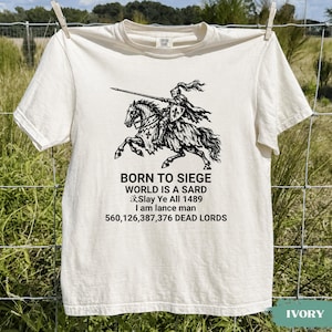 Puede incluir: Una camiseta de manga corta color marfil con un gráfico negro de un caballero a caballo sosteniendo una lanza. El texto dice: BORN TO SIEGE, WORLD IS A SARD, Slay Ye All 1489, I am lance man, 560,126,387,376 DEAD LORDS. Tiene cuello redondo.