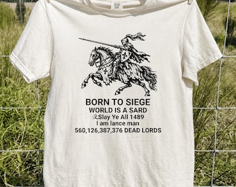 May include: An ivory short-sleeve t-shirt featuring a black graphic of a knight on horseback holding a lance. The text reads: BORN TO SIEGE, WORLD IS A SARD, Slay Ye All 1489, I am lance man, 560,126,387,376 DEAD LORDS. The shirt has a crew neckline.