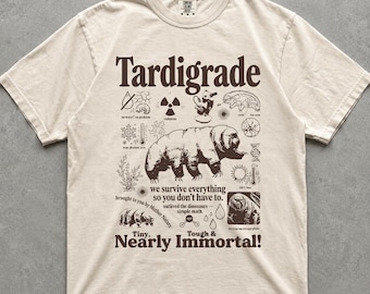 May include: A cream-colored short-sleeve t-shirt featuring a dark brown graphic print of a tardigrade surrounded by scientific icons and text including "Tardigrade," "we survive everything so you don't have to," and "Tiny, Nearly Immortal!"