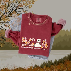 Puede incluir: Una sudadera granate con las letras "BCBA" en un diseño de hojas de otoño, con un gráfico de oso polar. El texto "BOARD CERTIFIED BEHAVIOR ANALYST" está debajo. La sudadera está doblada sobre unos vaqueros azul claro.