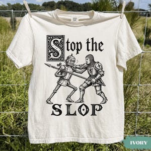 May include: An ivory short-sleeve t-shirt featuring a black graphic print of a medieval knight in chainmail armour fighting a robot with a spear. The text "Stop the SLOP" is printed in a decorative black font above and below the illustration.