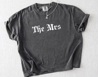 May include: A charcoal grey short-sleeve t-shirt with a faded wash, featuring the white text The Mrs in a gothic-style font across the chest. A thin silver-toned chain necklace with a small circular pendant rests on the neckline of the shirt.