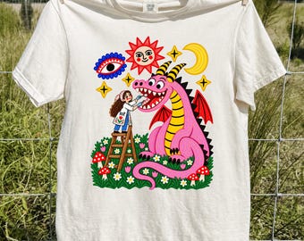 May include: An ivory short-sleeve t-shirt featuring a colorful graphic print of a person on a wooden ladder cleaning the teeth of a large pink dragon. The design includes a sun, moon, eye, stars, and red mushrooms in a grassy field setting.