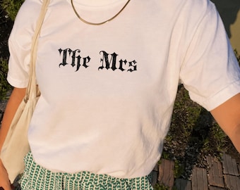 May include: A white short-sleeved t-shirt featuring the text The Mrs printed in a black gothic-style font across the chest. The shirt is paired with green and white patterned shorts and a light-colored tote bag, complemented by a gold chain necklace.