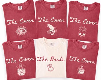 May include: Five red t-shirts and one white t-shirt. The red shirts feature the text The Coven with various white line art graphics including an eye, a cat, a skull, a potion bottle, and a skeleton. The white shirt says The Bride with a ring.