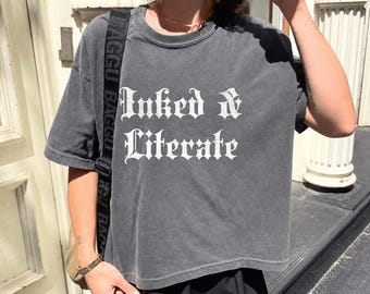 May include: A charcoal gray short-sleeve t-shirt featuring the white text Inked & Literate in a gothic-style font. The shirt has a crew neckline and a relaxed fit. A black bag strap with the repeating white text BAGGU is visible across the shoulder.