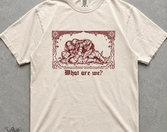May include: A cream-colored short-sleeve t-shirt featuring a dark red graphic print of two armored knights wrestling on the ground, enclosed in a decorative rectangular border. Below the illustration, the text reads, What are we? in a gothic font.
