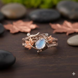Puede incluir: Un anillo de oro rosa con un diseño inspirado en la naturaleza. El anillo presenta una piedra preciosa azul claro en forma de pera, detalles de hojas de arce y pequeños diamantes. La banda está diseñada para parecer ramas entrelazadas.
