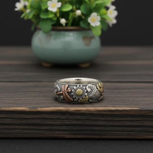 Puede incluir: Un anillo de plata con un diseño floral e insectos detallado. El anillo presenta una libélula, una margarita con un centro dorado y otros elementos florales. El anillo está sobre una superficie de madera oscura.