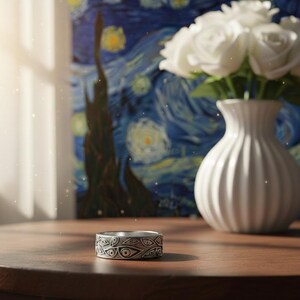 Op de afbeelding: Een zilveren ring met een oogpatroon ligt op een houten tafel. Een witte vaas met witte rozen en een schilderij van een sterrennacht zijn op de achtergrond. De ring, met zijn gedetailleerde ontwerp, is het centrale element.