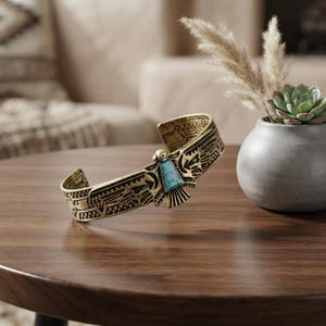 May include: A gold-toned metal cuff bracelet featuring an engraved bird motif with a triangular turquoise-coloured inlay at the centre. The band displays intricate geometric patterns along its entire length, showcasing a wide, open-ended design.