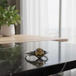 May include: A silver ring with a gold bee emblem. The ring features a detailed bee design in the center, surrounded by a circular frame. The ring is set against a black marble surface, with a blurred background.