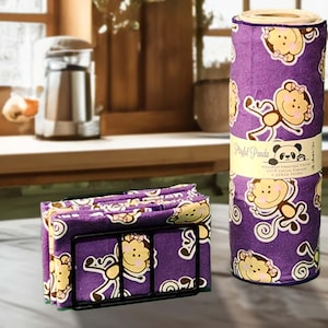 May include: A set of purple kitchen towels with a monkey pattern, displayed in a black metal holder and a roll. The roll has a label with the text "Bright Panda" and a panda logo. The towels are on a wooden surface.