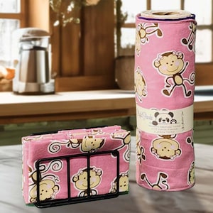 May include: Pink kitchen towels with a monkey pattern, displayed in a black metal napkin holder and a rolled-up cylinder. The towels feature a repeating design of smiling monkeys. A label on the rolled towels reads "Panda".