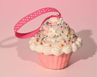 May include: A pink cupcake ornament with white frosting, colorful sprinkles, and a pink ribbon with white polka dots. The cupcake has a glittery pink base and is designed for hanging.