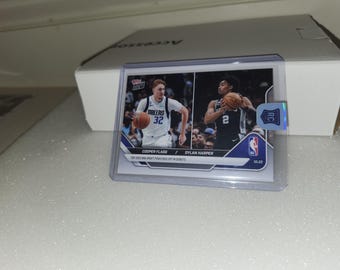 May include: A basketball trading card featuring Cooper Flagg and Dylan Harper. The card is encased in a protective sleeve. The card has the NBA logo and a Rookie Card emblem. The text on the card reads "TOP 2023 NBA DRAFT PICKS FACE OFF IN DEBUTS".