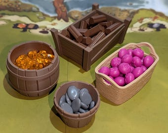 May include: A tabletop game setup with various resource tokens. Includes a wooden cart filled with brown logs, a barrel of amber-colored gems, a basket of pink star-shaped fruits, and a small bowl of gray stones. The game board is visible in the background.
