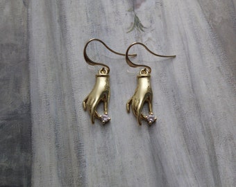 May include: A pair of gold dangle earrings featuring a hand holding a small clear rhinestone. The earrings have a simple design and are perfect for everyday wear.
