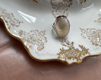 May include: Oval-shaped mother-of-pearl ring with a silver band. The ring sits on a white porcelain dish with gold filigree and trim. The dish has a curved shape and is resting on a pink surface.