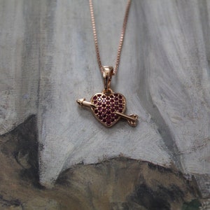 Może przedstawiać: Złoty naszyjnik z różowego złota z sercem w kształcie wisiora. Serce jest pokryte małymi czerwonymi kamieniami szlachetnymi i ma strzałę przechodzącą przez nie.