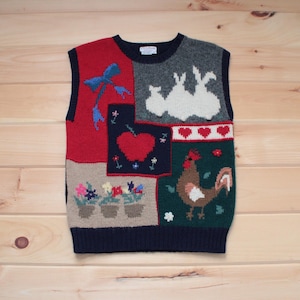 Könnte beinhalten: Eine ärmellose Strickweste mit Patchwork-Design, das einen roten Apfel, einen Hahn, ein Kaninchenpaar und eine Schleife zeigt. Die Weste ist aus einer weichen, warmen Wollmischung gefertigt und hat einen klassischen, zeitlosen Stil.