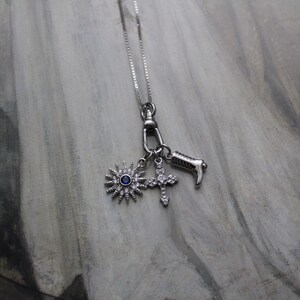 Puede incluir: Un collar de plata con un dije que presenta una piedra preciosa azul, una estrella de rayos y una cruz. Los dijes están unidos a una cadena de plata.