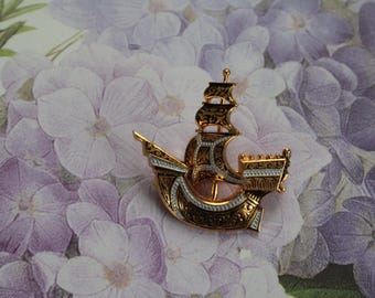 May include: A gold and white ship brooch with intricate details. The ship has a tall mast and sails, and the hull is decorated with a floral pattern.