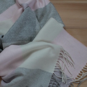 May include: A rectangular scarf in grey, pink and off-white. The scarf has a fringed edge and a colour-block design. The scarf is made of a soft material and is suitable for cooler weather.