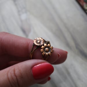 May include: A gold-toned ring with two delicate flower designs. The ring is worn on a finger with red nail polish.