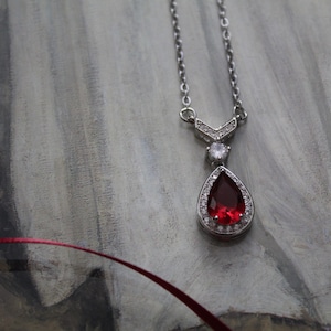 Puede incluir: Un collar de plata con un colgante de piedra roja en forma de lágrima rodeado de piedras transparentes más pequeñas.