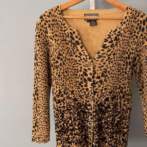 Könnte beinhalten: Beigefarbene Strickjacke mit Leopardenmuster und langen Ärmeln. Der Cardigan mit Knopfleiste hat unten schwarze Paillettenverzierungen. Das Kleidungsstück hängt an einem Holzbügel.