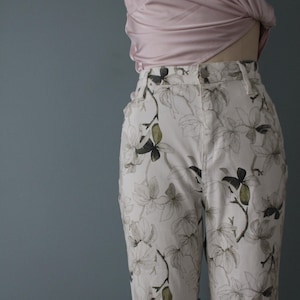 Może przedstawiać: Białe spodnie z kwiatowym wzorem z szarymi i zielonymi liśćmi i kwiatami. Spodnie są z wysokim stanem i mają zapięcie na guzik i zamek błyskawiczny. Widoczny jest również jasnoróżowy top z koronkowymi detalami.
