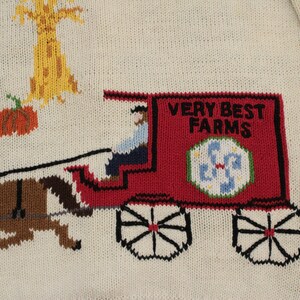 May include: A white knitted sweater with a red wagon pulled by two brown horses. The wagon has a sign that says "Very Best Farms". The sweater also has a small tree and pumpkins.