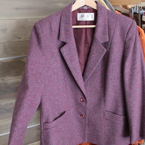 May include: A purple blazer with a two-button closure and two front pockets. The blazer is hanging on a wooden hanger. The jacket has a classic lapel and a burgundy lining. The fabric has a textured appearance.