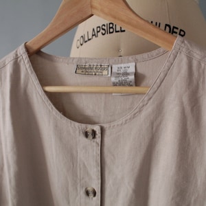 May include: A beige button-down shirt with a scoop neckline, hanging on a wooden hanger. The shirt has a label that reads "STEPHANIE ROGERS by Fashion Designs". The shirt is made of a light, textured fabric and has two visible buttons.