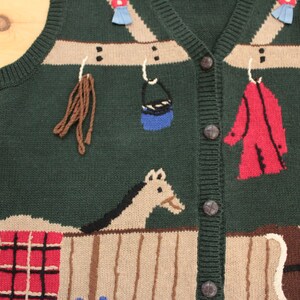 Puede incluir: Un chaleco de punto verde con un diseño estampado en marrón, rojo y azul. El chaleco presenta un caballo, un cubo y otros artículos colgados de una barra de madera.