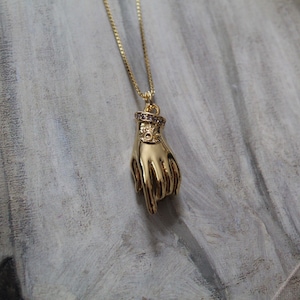 May include: Gold-colored pendant necklace featuring a hand pointing downward. The hand pendant has a ring with small purple stones. The necklace hangs on a delicate gold chain. The background is a textured, neutral-toned surface.