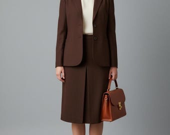 May include: A dark brown blazer and matching knee-length skirt with a centre pleat, worn over a cream-coloured top. The outfit is paired with black Mary Jane shoes and a structured tan leather handbag with a gold-tone clasp and top handle.