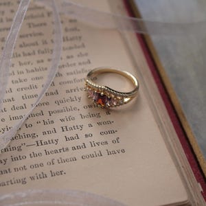 May include: A gold ring with a split band, adorned with a central red gemstone and smaller clear stones. The ring rests on an open book with aged pages and a white ribbon.